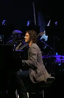 Josh Groban est assis devant son piano, devant ses musiciens.