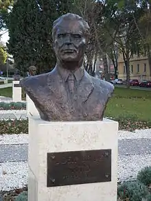 Photo couleur d'un buste de Josip Broz Tito de couleur sombre, posé sur un support cubique en pierre blanche.