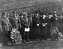 Photographie noir et blanc de trois hommes en habit cravate entourés de nombreux soldats en uniforme.