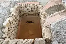 Vue d'une cave aux murs appareillés creusée dans le sol d'une cour antique ; deux canalisations en tuiles aboutissent à la cave.