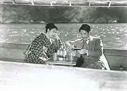 Une femme en kimono (Kinuyo Tanaka) et un homme en costume (Sō Yamamura) sont assis dans un bateau. Entre eux se dresse une petite table sur laquelle deux verres et une bouteille sont posés. La femme a le coude appuyé sur le bord du bateau tandis que l'homme tient un carnet et un stylo (image en noir et blanc).