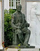 Statue représentant un homme assis portant barbe et moustache, tenant un livre dans sa main.