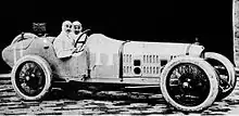 Photo de deux hommes en combinaison de pilote blanche, prenant la pose dans le poste de pilotage d'une voiture.