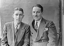 Photographie en noir et blanc de deux hommes en costumes assis côte à côte.