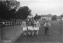 Photographie en noir et blanc de quatre hommes portant en triomphe un athlète au milieu d'un stade plein.