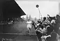 Photographie en noir et blanc d'un coureur à pied à l’arrivée d'une course, encouragé par des hommes qui crient.