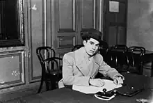 Photographie en noir et blanc d'un homme portant un chapeau signant un document à un bureau.