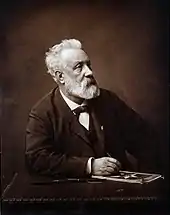 Photo noir et blanc de Jules Verne, un homme barbu aux cheveux blancs assis devant une table, le crayon à la main.