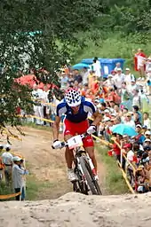  Homme debout sur les pédales d'un VTT portant une tenue bleu-blanc-rouge avec un public en arrière-plan.