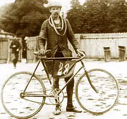 Photographie en noir et blanc d'un homme moustachu tenant son vélo, avec un tissu portant le no 28 au milieu du cadre.