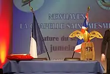 Photographie d'une estrade de part et d'autre de laquelle peuvent être vus le drapeau français et le drapeau du Royaume-Uni et en arrière plan le nom des deux communes jumelées des deux pays.