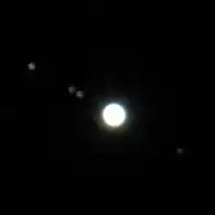 Jupiter apparait très brillante devant le fond noir. Les lunes apparaissent comme quatre petits points gris et flous.