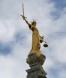 Une statue dorée d'une femme couronnée, tenant un glaive et une balance, se détache sur fond de ciel nuageux.