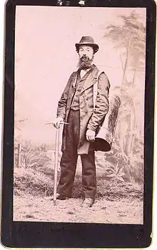 Photographie noir et blanc d'un homme debout, appuyé sur un piolet et portant en bandoulière une grande boîte cylindrique en fer-blanc