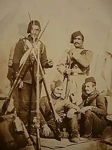 Photographie de quatre soldats moustachus portant des fez. Deux sont assis à côté d'un trépied de fusils équipés de baïonnettes.