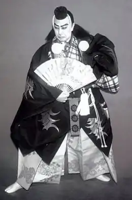 Photographie en noir et blanc d’un acteur de théâtre kabuki habillé en kimono. Il a le visage maquillé de blanc, avec des sourcils épais et une barbe noire. Il tient un éventail dans sa main droite et regarde vers la gauche avec une expression féroce.