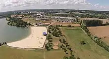 Photographie aérienne d'une base de loisirs comprenant un lac, une plage aménagée et des espaces verts.