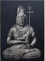 Front view of a statue in lotus position. The palm of the right hand is turned upward and held in front of the stomach. The left hand, close to the right foot, is holding a long pole with decorations at the end resembling a trident.