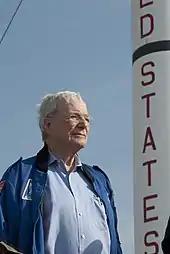 Un homme âgé, debout à côté d'une fusée visible partiellement.