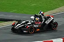 Sébastien Loeb avec casque et combinaison, au volant d'un KTM X-Bow avec un passager, sur une piste goudronnée.
