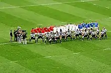 Vue de loin de deux équipes alignées de part et d'autre de la ligne centrale, les Français, de face, portant de la gauche vers la droite des survetements rouge, blanc, bleu.