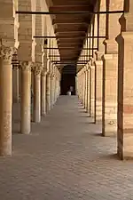 Vue intérieure montrant une double rangée d'arcades en perspective dans le portique précédant la salle de prière. Les arcs sont de type outrepassé (brisé ou en plein cintre).