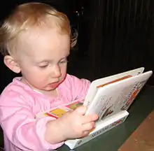 Un bébé est assis sur le sol. Il regarde un livre qu'il tient entre ses mains avec une expression d'intérêt et de concentration.