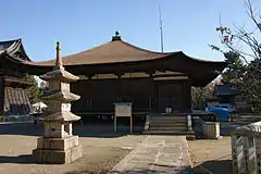 Wooden building with a pyramid shaped roof and an open veranda on the fron. A stair leads at one end of the veranda to the building's level.