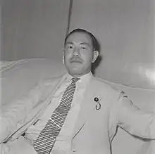 Photo noir et blanc d'un homme en costume clair, assis sur un canapé.