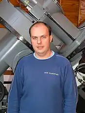 Half-length portrait of a man with descending brown hair. He is standing, looking into the camera and smiling. He is wearing a blue sweatshirt. A part of an astronomical telescope is visible in the background.