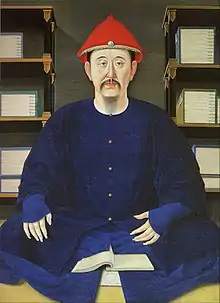 Très grande sobriété de décor et de costume. Assis en tailleur, de face, chapeau pointu rouge. Costume bleu. Un livre ou un cahier ouvert devant lui.