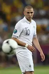 Photo de Karim Benzema vêtu du maillot blanc de l'équipe de France, regardant un ballon en l'air au premier plan.