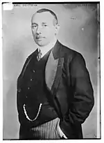 Photo en noir et blanc de profil d'un homme en costume, il porte une petite moustache et des cheveux courts