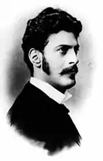 Photographie en noir et blanc du visage d'un homme aux cheveux bruns, portant moustaches et favoris, de profil droit.