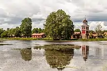 Un lac avec quelques nénuphars. De beaux arbres sur la berge, sous lesquels quelques bâtiments rouges se blottissent. Une petite tour donne l'heure.