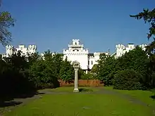 Photographie en couleurs d'un imposant château de style Tudor et aux façades blanches, flanquées de tourelles derrière un jardin planté de conifères vert tendre.