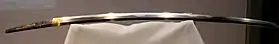 Photo couleur de la lame d'un sabre sur un portoir blanc (fond brun).