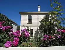 Une maison blanche de style colonial est entourée de verdure et de roses violacées au premier plan.