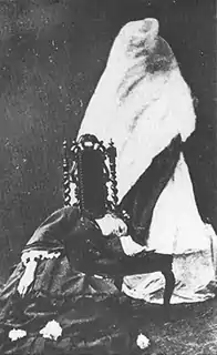 photographie noir et blanc : une jeune femme en noir endormie sur une chaise ; une forme dans un drap au dessus d'elle