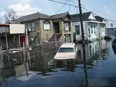 Photographie d'une rue inondée avec une voiture dans l'eau et plusieurs maisons aux fondations recouvertes.