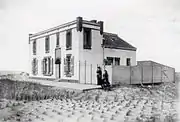 Photographie en noir et blanc d'une bâtisse et de ses résidents, au bord d'une plage.