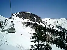 Photo couleur d'une remontée mécanique (à gauche), au-dessus des pentes enneigées et boisées d'un massif de montagne, sous un ciel bleu sans nuages.