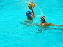 Deux joueurs de water-polo, l'un avec un casque bleu tient la balle dans sa main, l’autre défend avec un casque rouge et blanc.