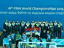 Une équipe sur un podium tenant un drapeau serbe, médailles autour du cou, devant des photographes accrédités.
