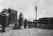 Photographie en noir et blanc montrant un portail d'entrée et un bâtiment en briques.