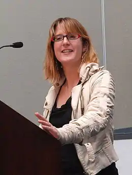 Une femme blonde portant des lunettes donne une présentation depuis un pupitre.