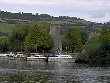 Vue de bateaux amarrés depuis la rive d'en face. Au-delà, se trouve une cheminée en pierre entourée d'arbres, avec des collines dans le lointain.