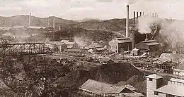Photographie d'une usine sidérurgique vue de loin. De longues cheminées dépassent d'un corps de bâtiments, et de la vapeur s'échappe de plusieurs endroits.
