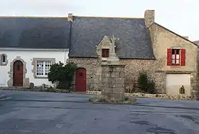 Photographie de trois maisons basses couvertes d’ardoise derrière une croix de granit.