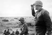Photo en noir et blanc d'hommes en uniforme avec au second plan des chars de combat.
